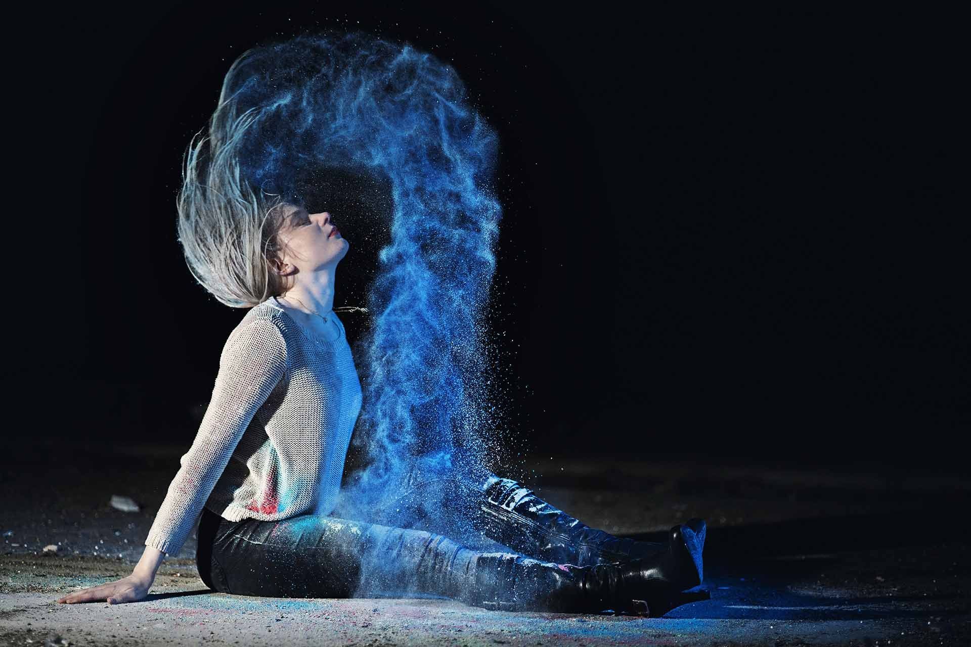 Eine Frau sitzt auf dem Boden und aus ihren Haaren kommt blaues Pulver.