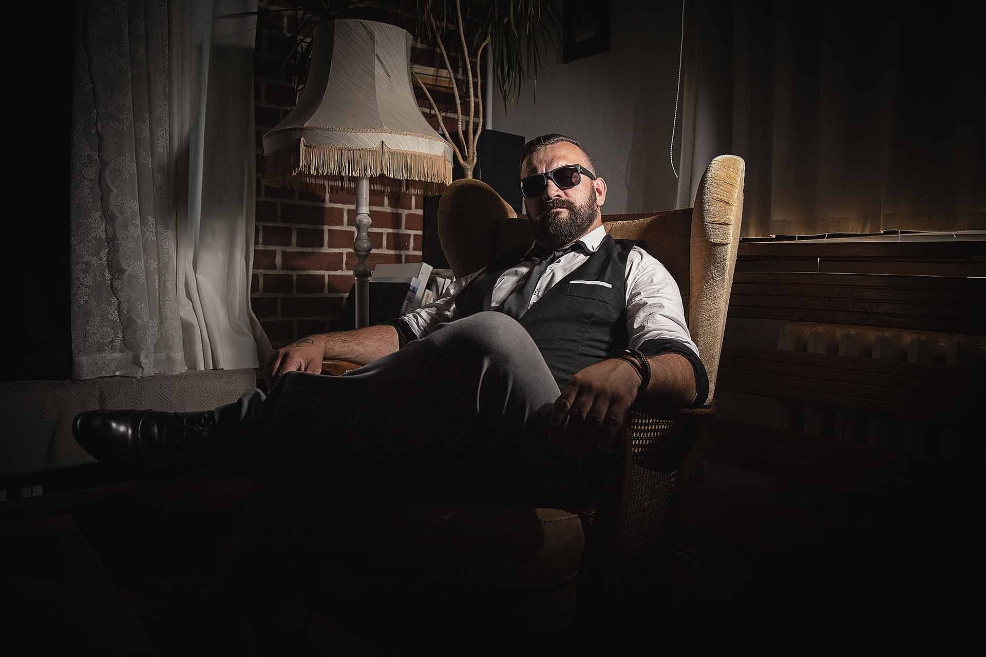 Ein Mann mit Bart sitzt in einem dunklen Raum auf einem Stuhl.