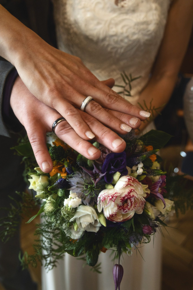 Eine Braut und ein Bräutigam stecken sich ihre Eheringe an die Hände und halten dabei einen Blumenstrauß in der Hand.
