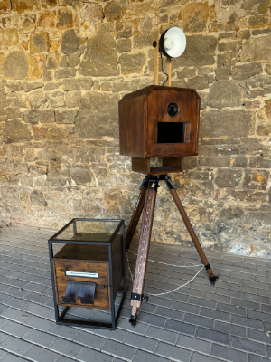 Eine hölzerne Fotokabine steht auf einem Stativ vor einer Backsteinmauer.