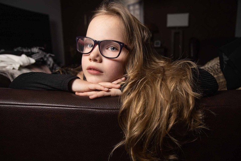 Ein Mädchen mit Brille ruht auf einem braunen Sofa und schaut in die Kamera.