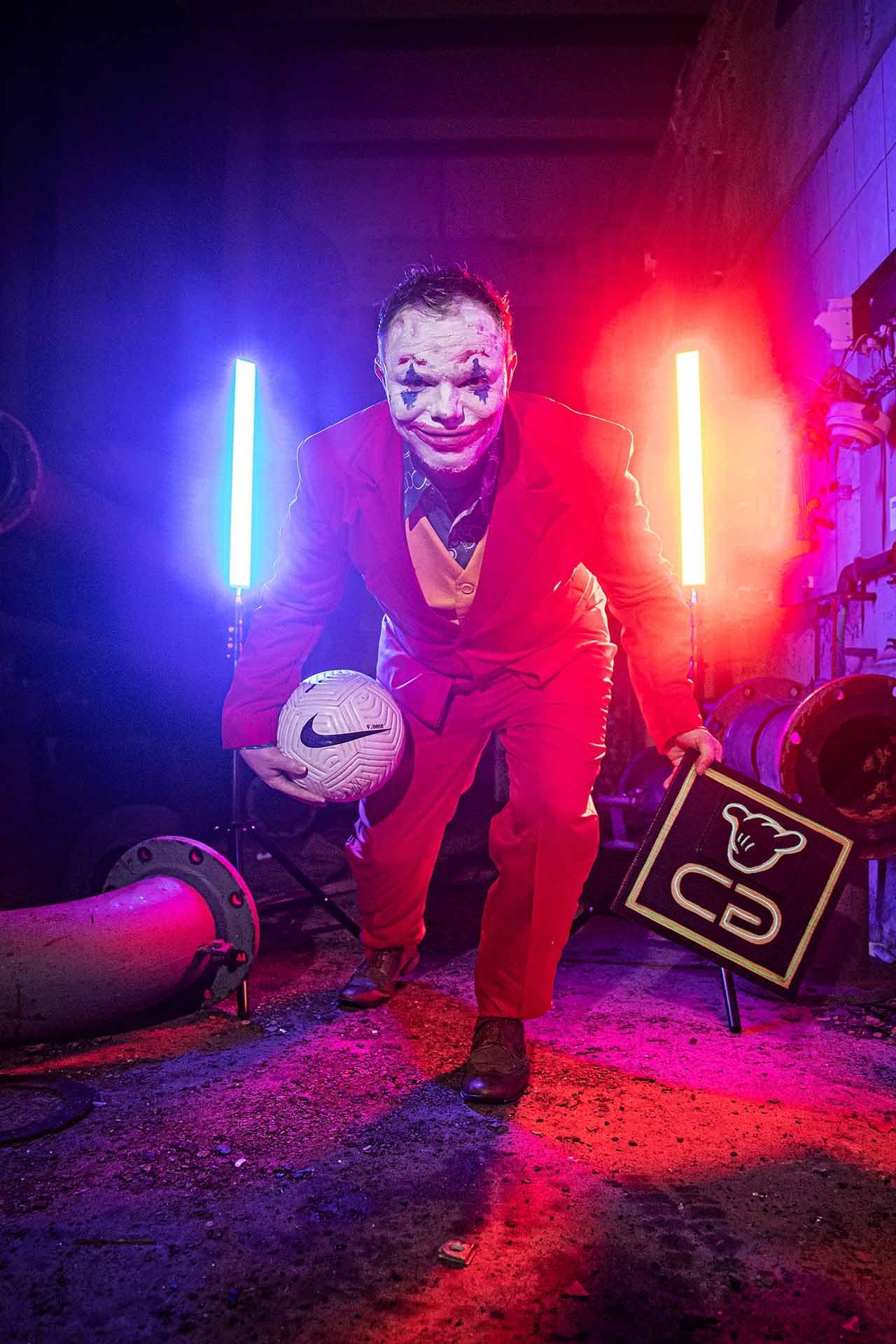 Ein als Joker verkleideter Mann hält in einem dunklen Raum einen Fußball in der Hand.