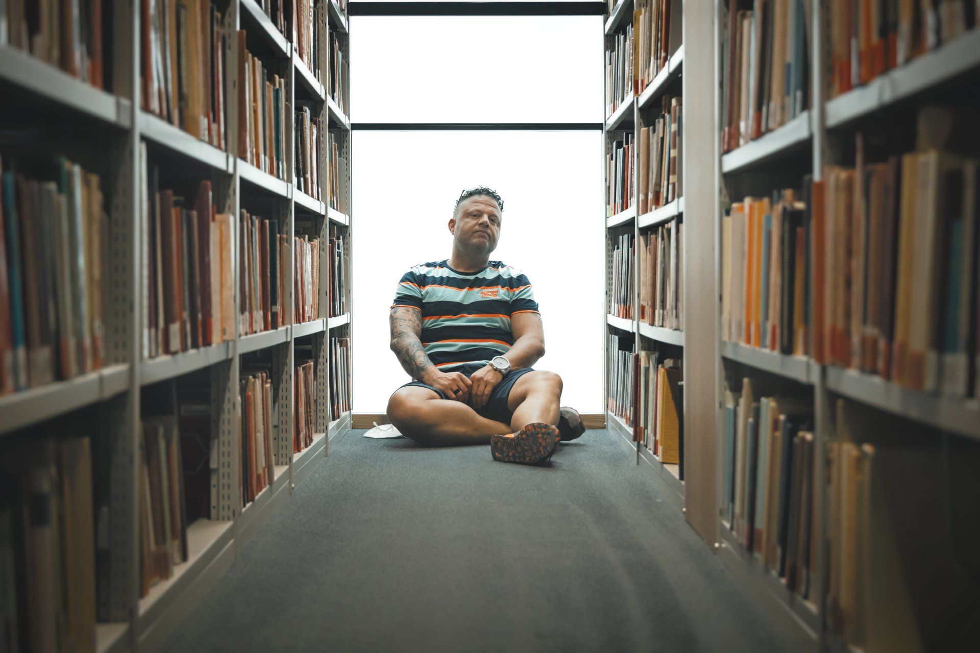 Ein Mann sitzt in einer Bibliothek auf dem Boden, umgeben von Bücherregalen.