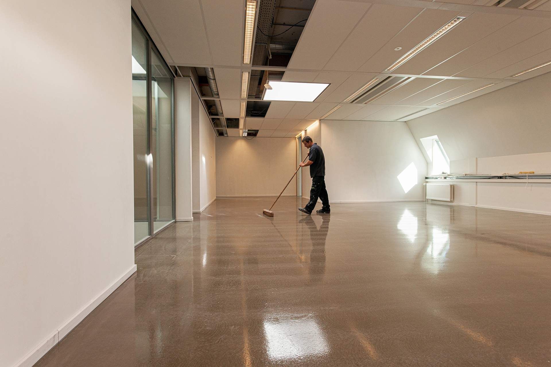 A person sweeps a shiny, empty room with white walls and a long window.