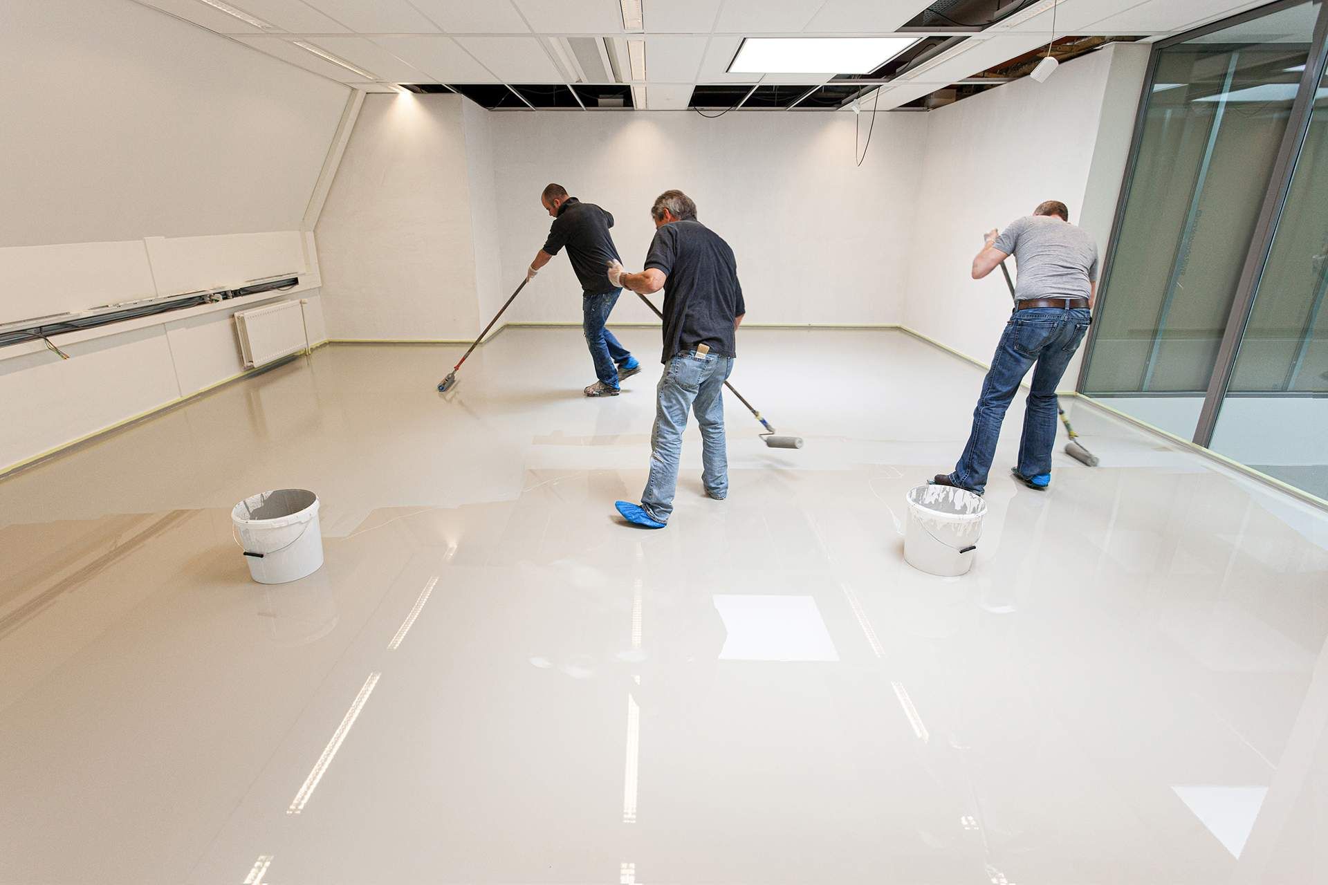 Three men applying epoxy flooring to a white room using long handled tools.