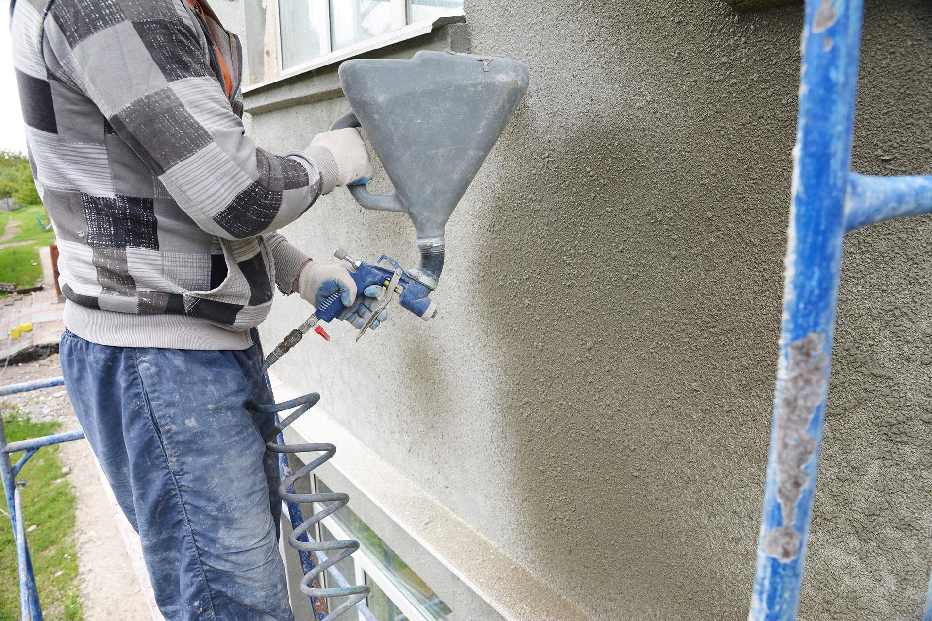 Person applying stucco to a building's exterior with a hopper gun, standing on a scaffold.