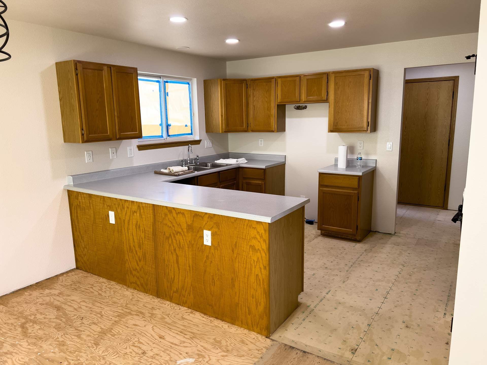 Kitchen with light-brown cabinets, gray countertops, and unfinished flooring. A doorway is visible on the right.