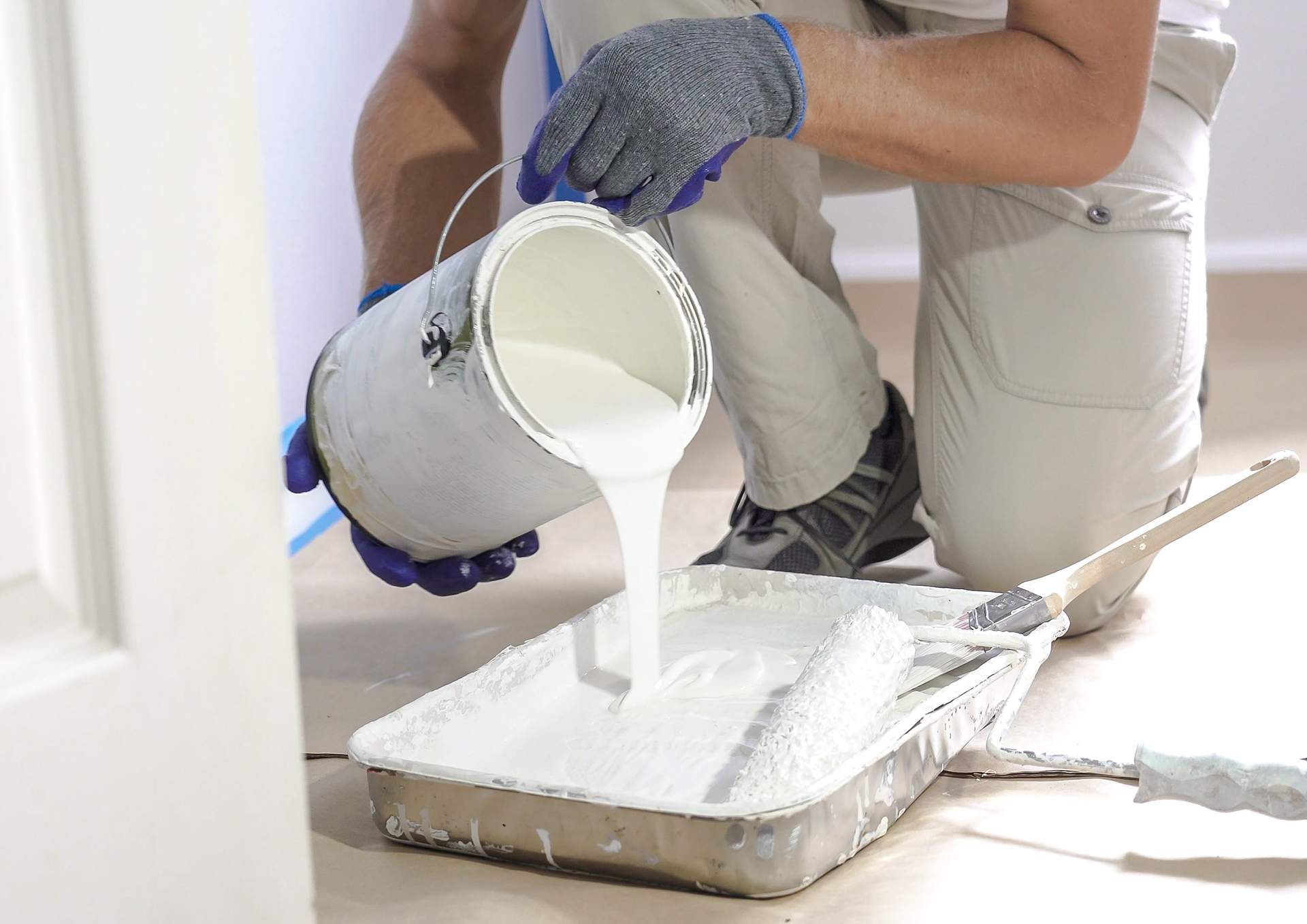 Painter pours white paint from a can into a tray; wearing gloves in a room.