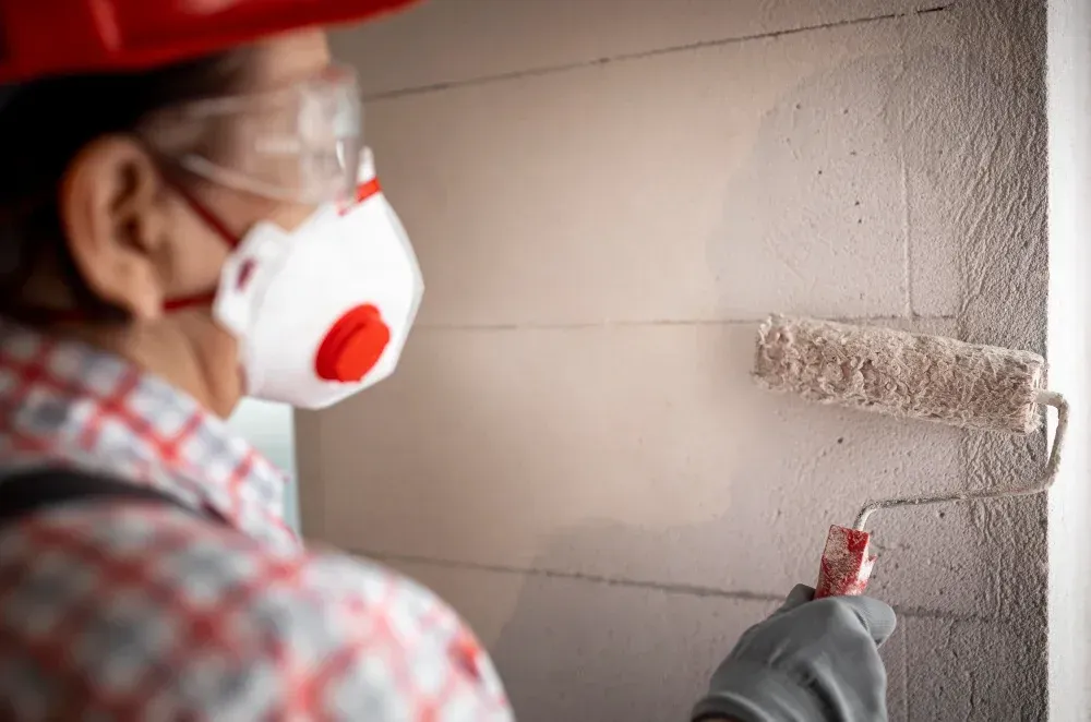 A man wearing a mask and safety glasses is painting a wall with a paint roller.