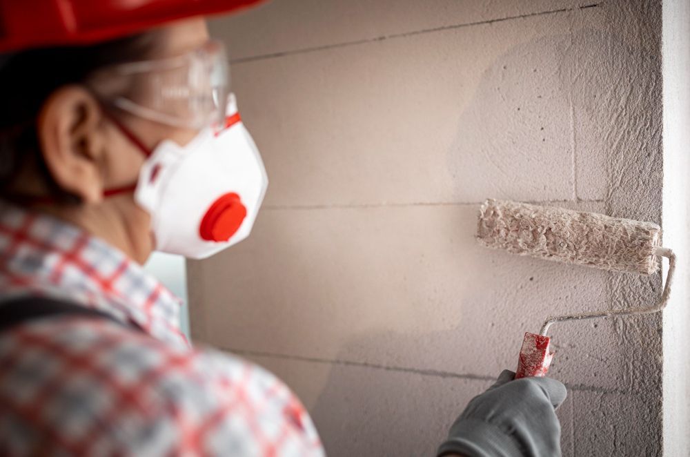 A man wearing a mask and safety glasses is painting a wall with a paint roller.