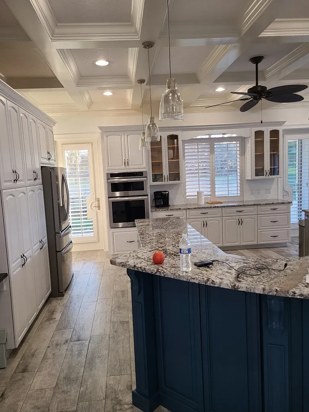 A kitchen with white cabinets , granite counter tops , a blue island and a ceiling fan.