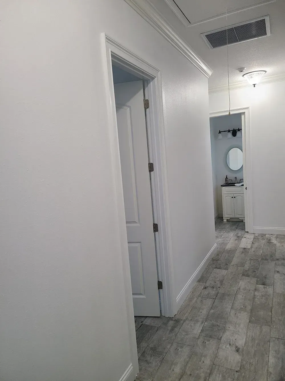A hallway with white walls and a wooden floor leading to a bathroom.