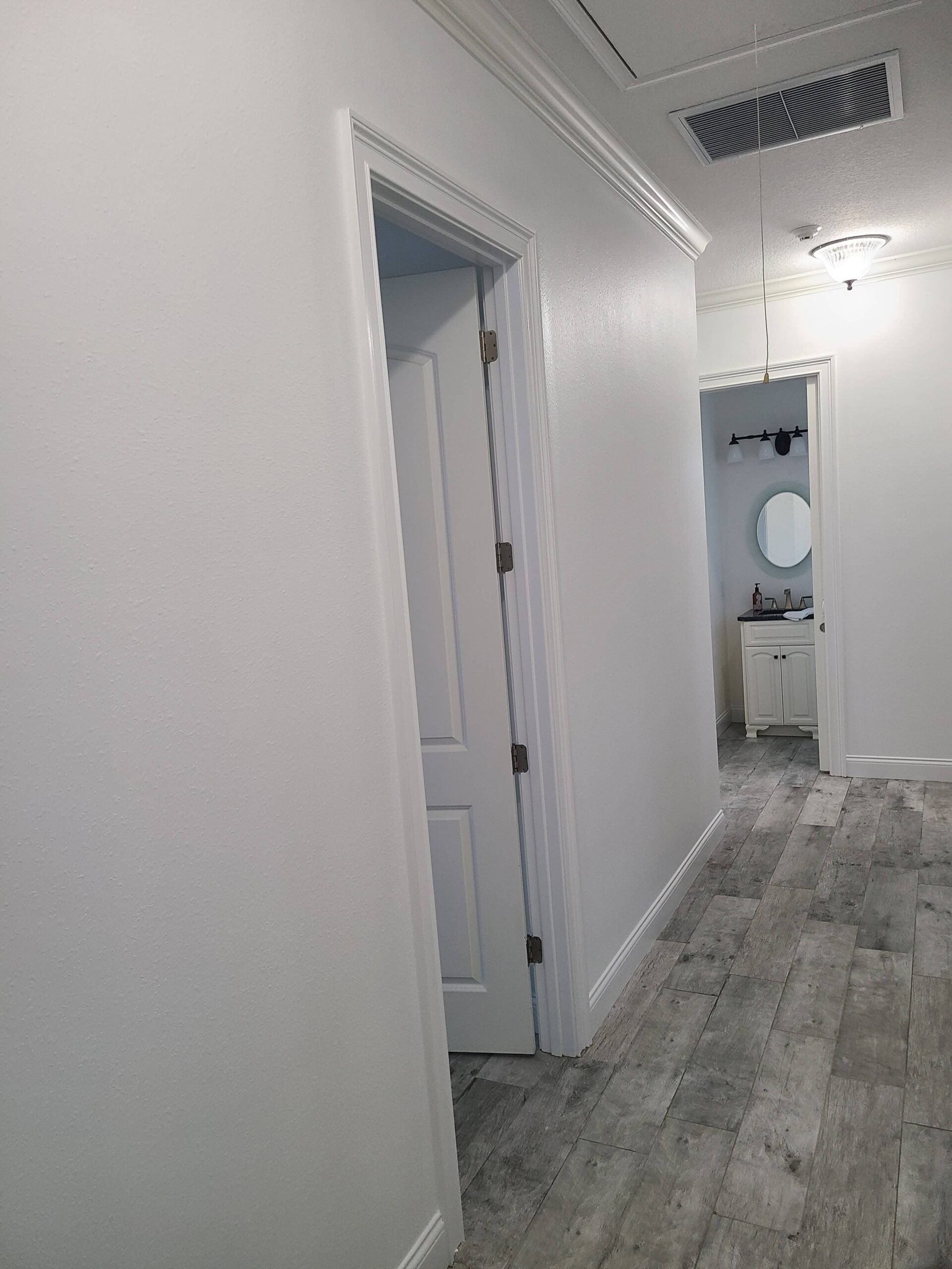 A hallway with white walls and a wooden floor leading to a bathroom.