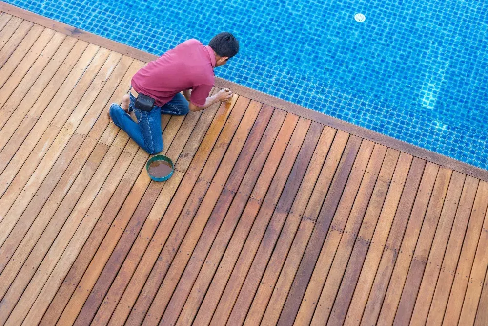 A man is kneeling on a wooden deck next to a swimming pool.