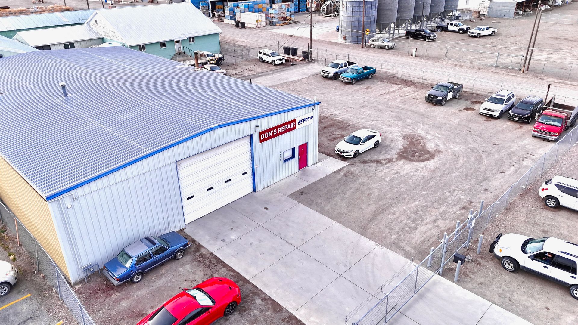 Aerial view of a metal building with a white garage door, several cars, and silos | Don's Repair