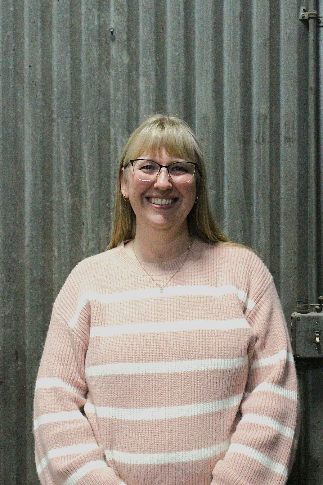 Woman with glasses and blonde hair smiles in a pink and white striped sweater in front of a corrugated metal wall | Don's Repair