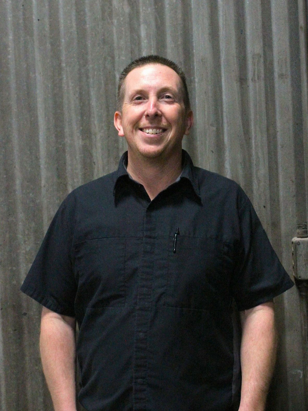 Man smiling in a dark blue short-sleeved shirt standing in front of a corrugated metal wall | Don's Repair