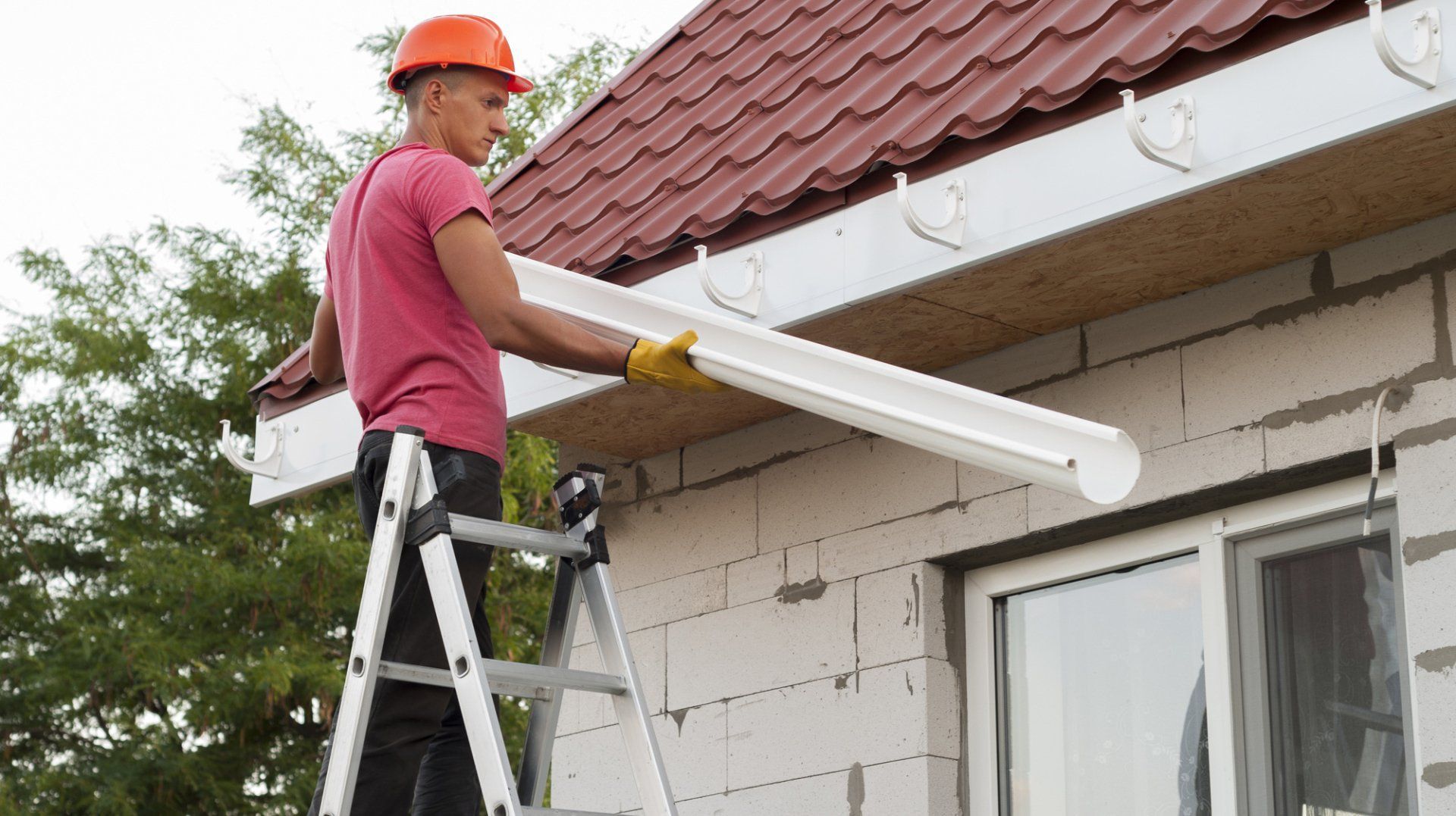 A man is standing on a ladder installing a gutter on the side of a house.