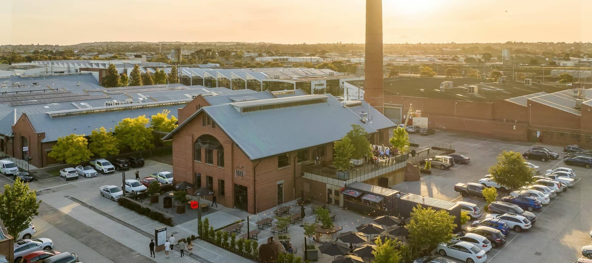 Aerial view of brick building with a tall chimney at sunset, surrounded by parking lots and other buildings.