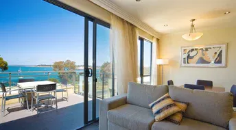 Living room with balcony overlooking a blue ocean; gray sofa, dining table, and sliding glass doors.