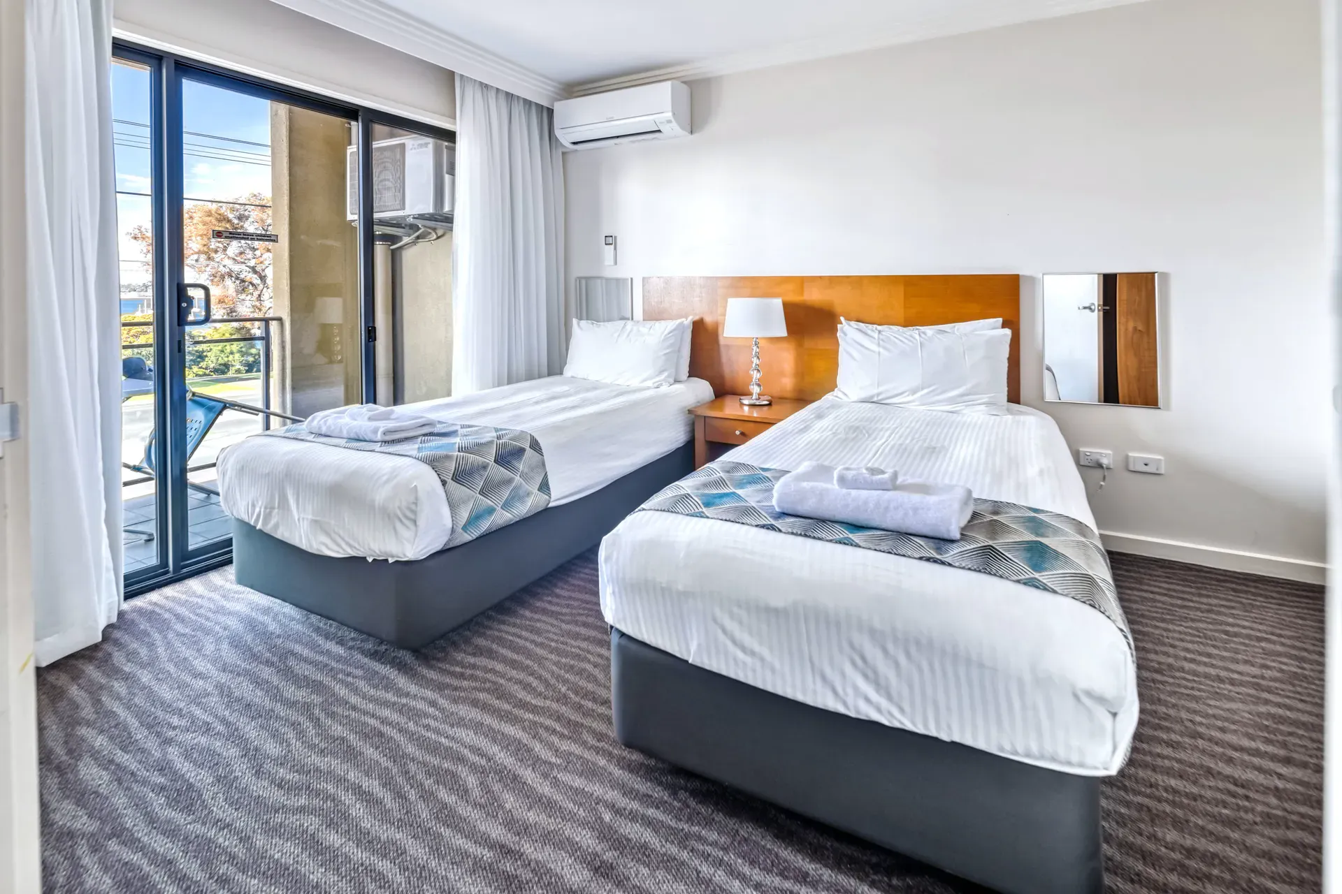 A hotel room with two twin beds, a small table, and a sliding glass door to a balcony.