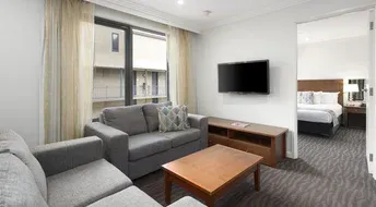 Modern hotel suite living room with gray couch, TV, and bedroom doorway.
