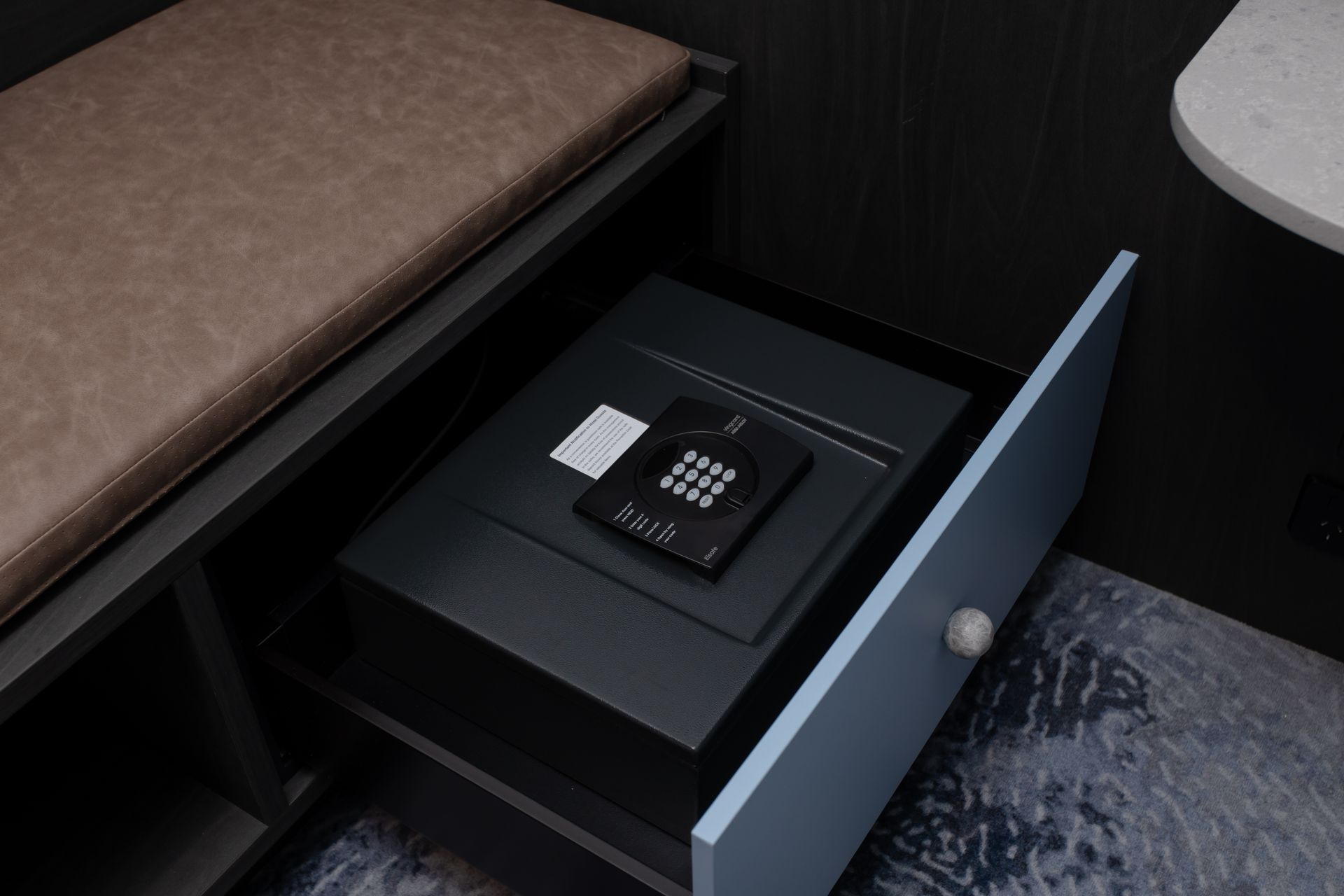 A partially opened drawer with a digital safe inside, under a brown bench in a hotel room.