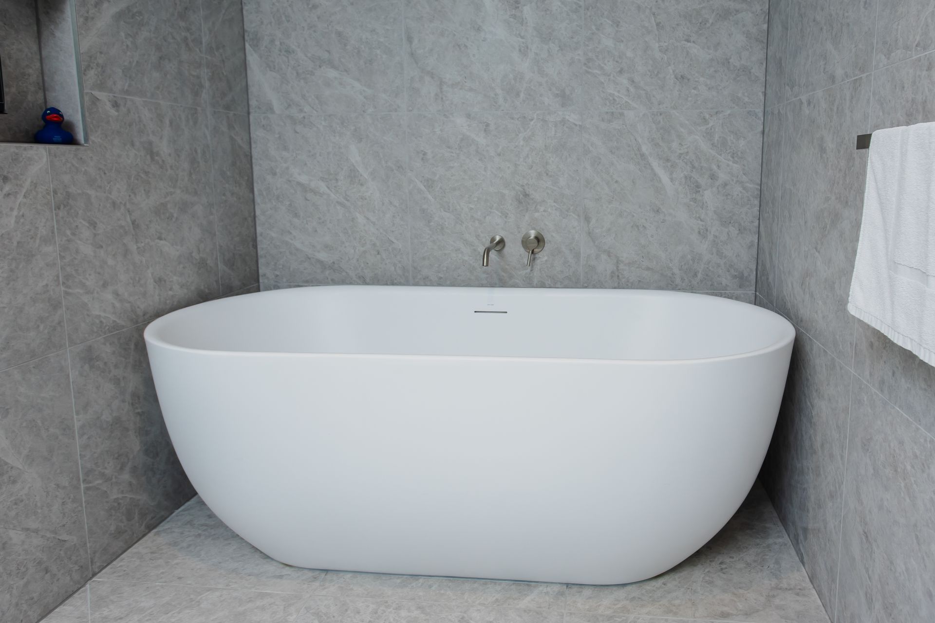 White oval bathtub in a gray-tiled bathroom. A towel hangs to the right. Faucet on the wall above.