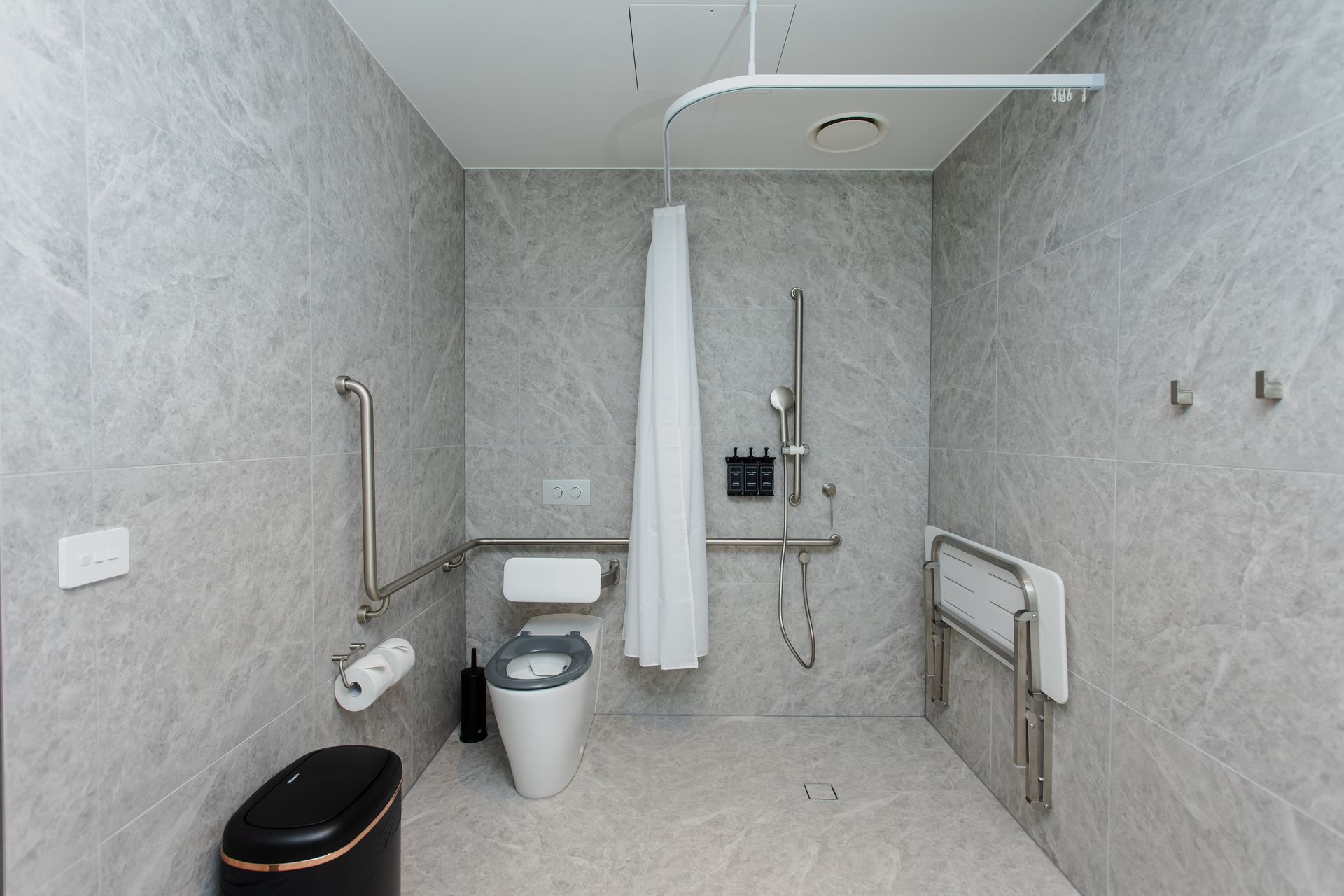 Accessible bathroom with gray tile walls, toilet, grab bars, and fold-down seat.