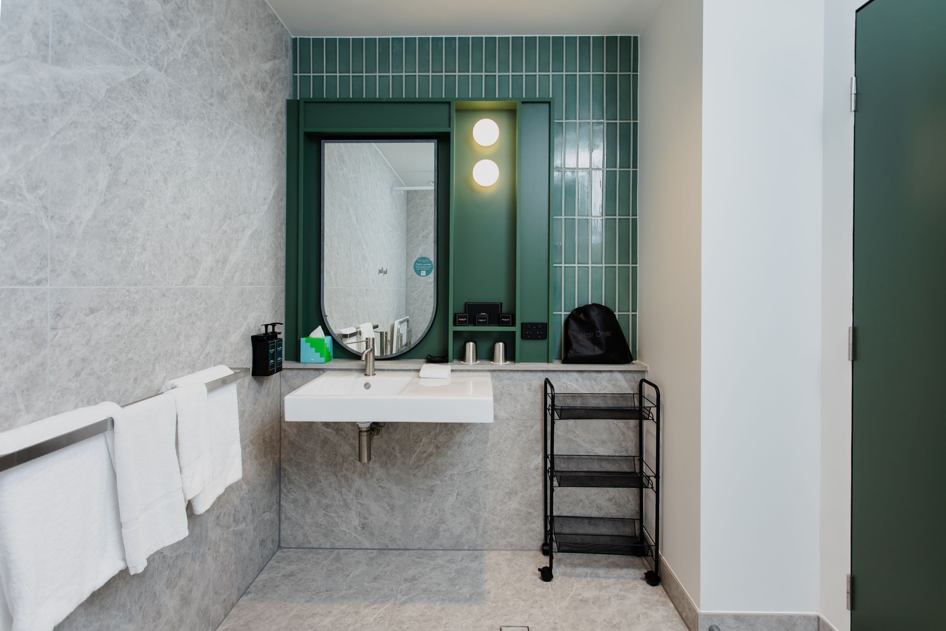 Bathroom with marble tile, a green-framed mirror, and a rolling black cart. White towels hang on a bar.