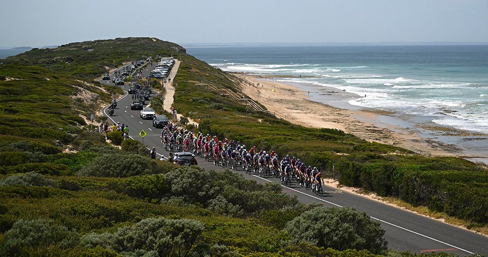 Cyclists race on a coastal road, ocean and beach to the right, lush green vegetation and a hilly landscape.