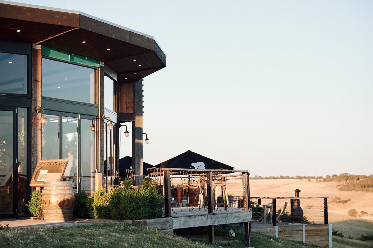 Modern building with large windows and deck overlooking a rural landscape at sunset.