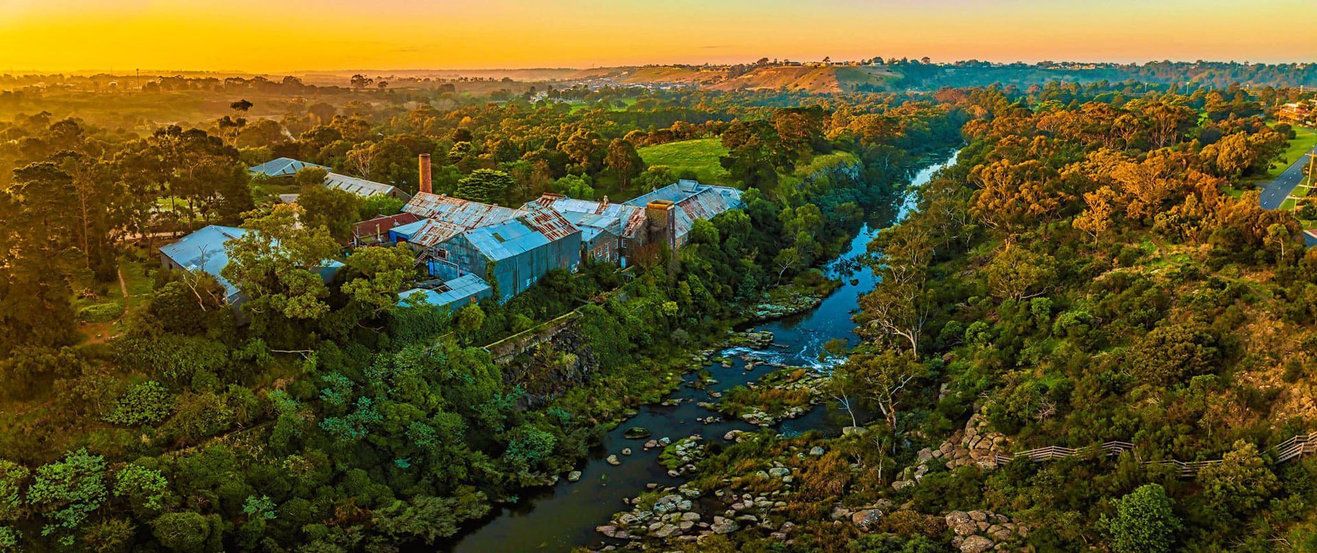 Aerial view of a river winding through a lush green forest, with buildings visible. Golden sunset.