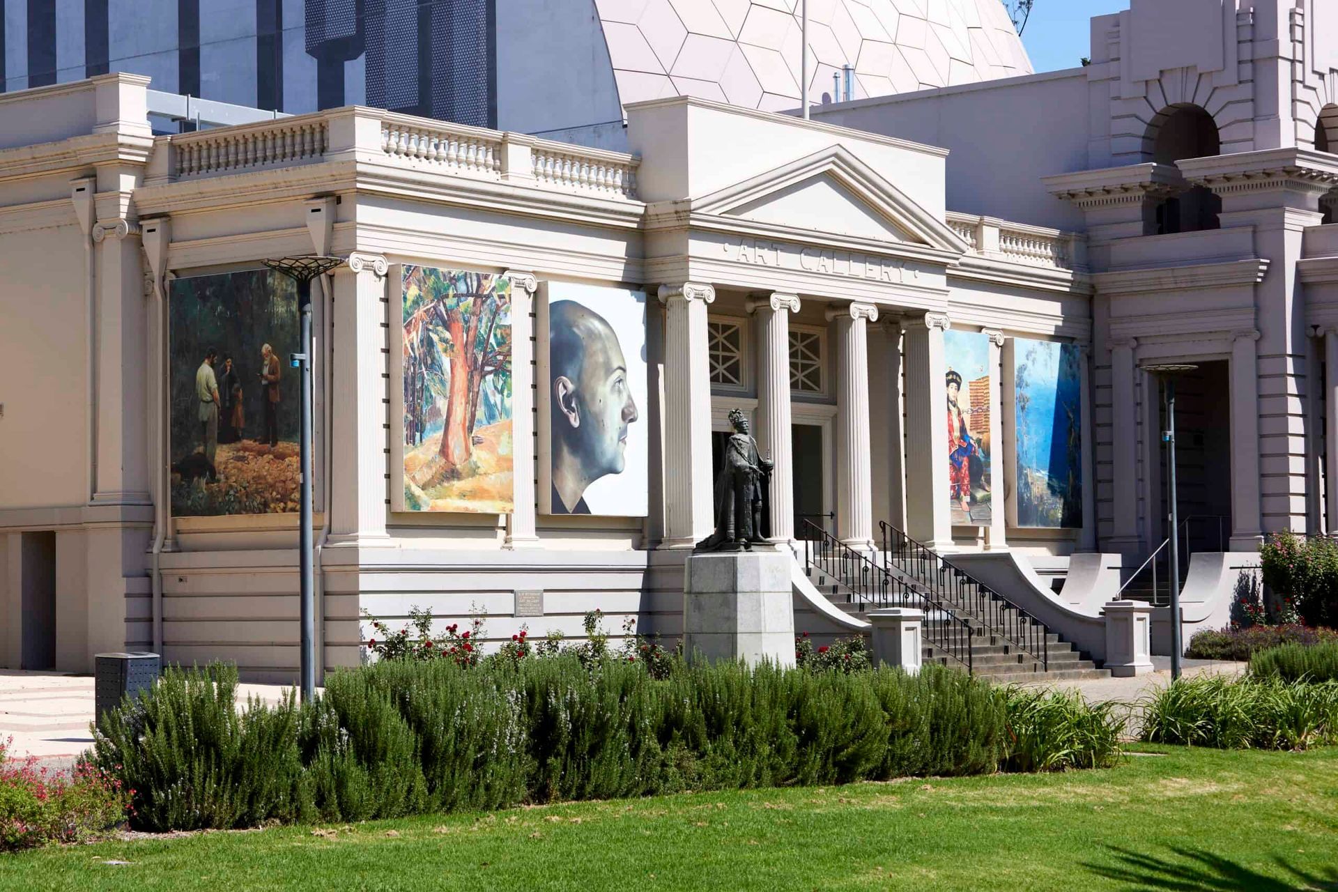 Neo-classical building with large art posters displayed; statue of a person in front; green grass.