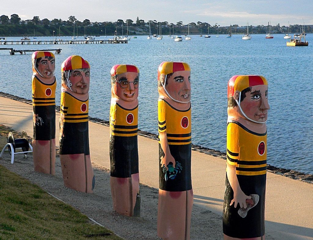 Five wooden lifeguard statues, yellow/black outfits, beach setting, sailboats in background.