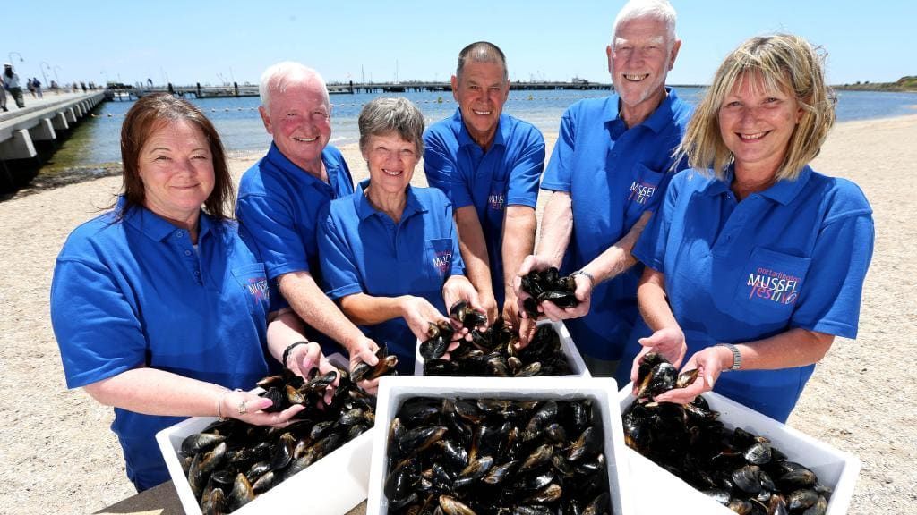 Group of six people holding mussels on a beach, wearing blue shirts with a logo.