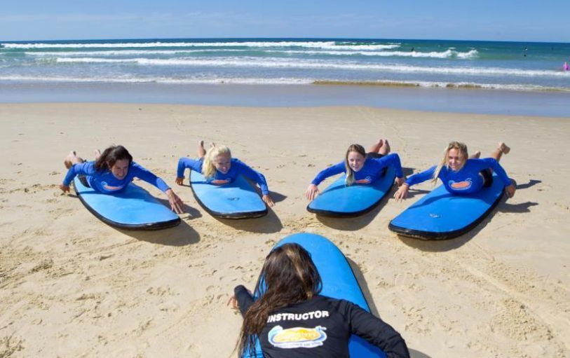 Surfing lesson: Instructor demonstrating paddling on a surfboard to students on a sunny beach.