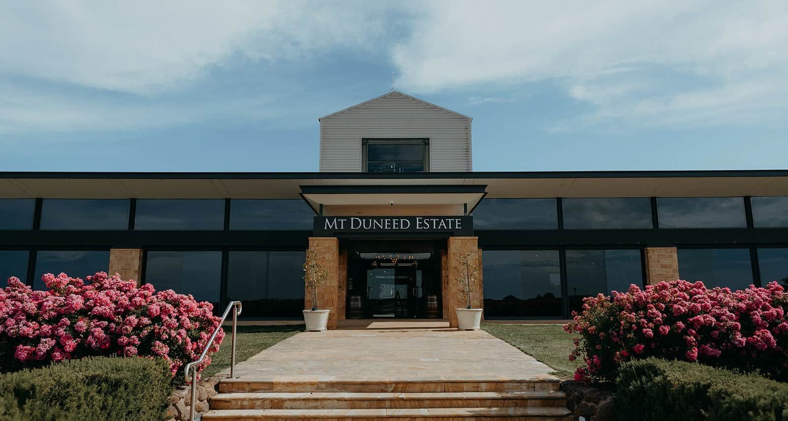 Modern building with glass windows, entrance framed by stone, and pink flowering bushes.