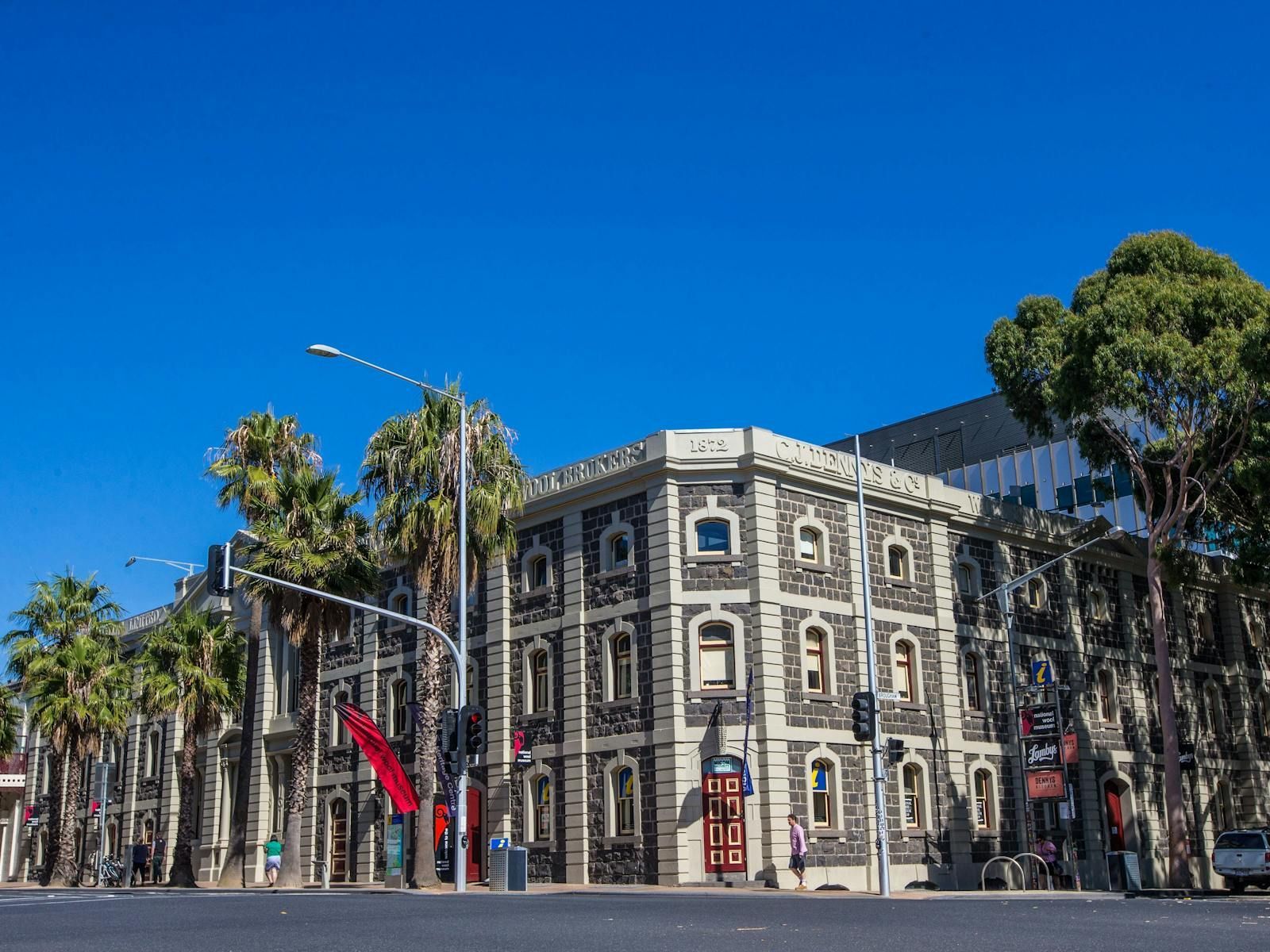 A historic building with dark stone facade, palm trees, and a bright blue sky in Melbourne, Australia.