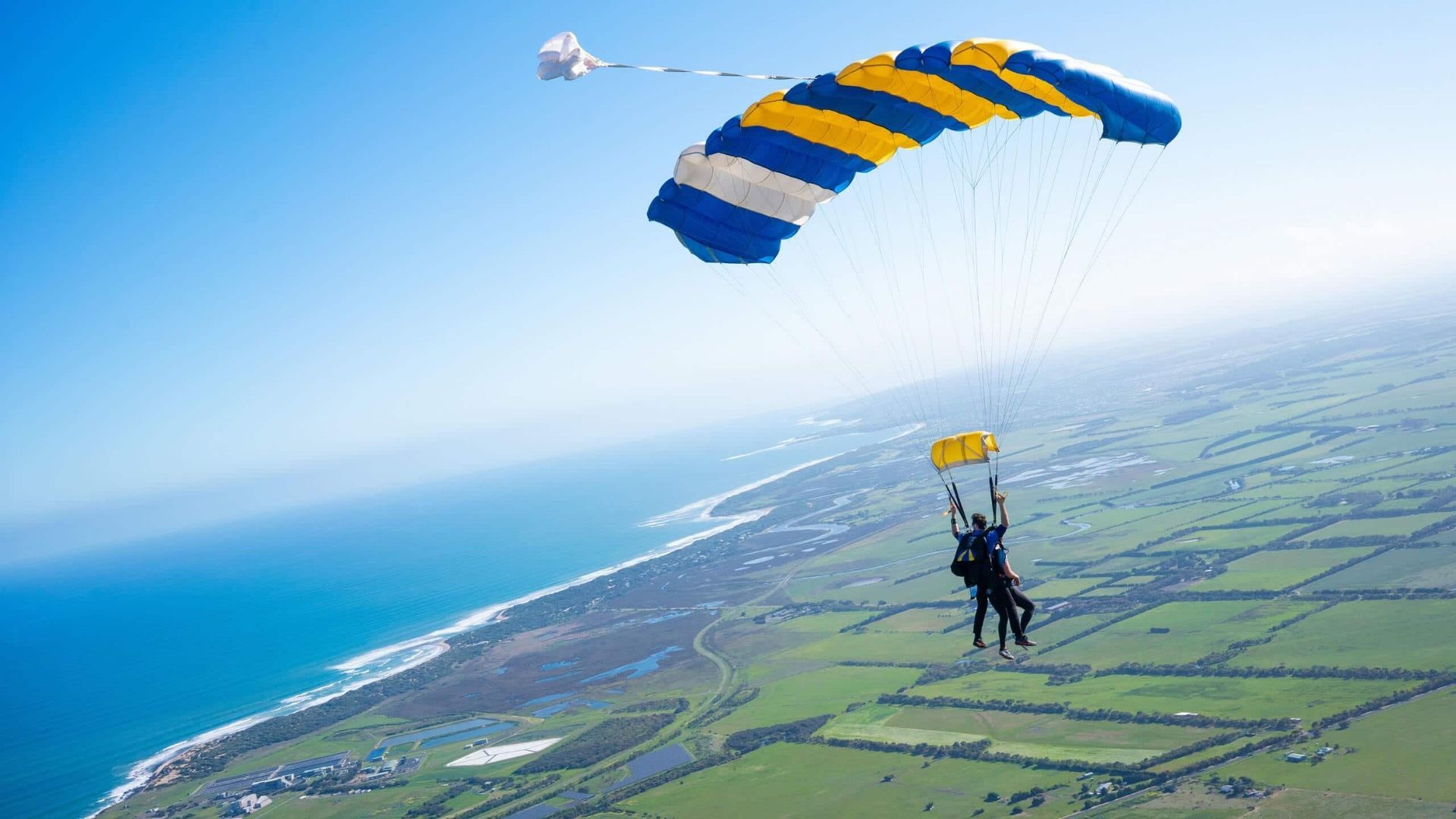 Skydivers under a colorful parachute over a green landscape and the ocean.