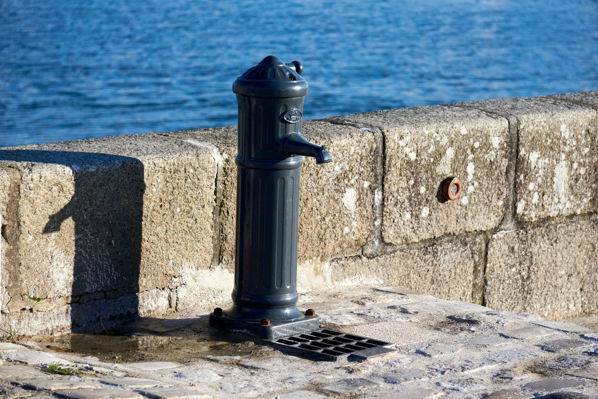 Black water fountain on a stone seawall beside blue water.