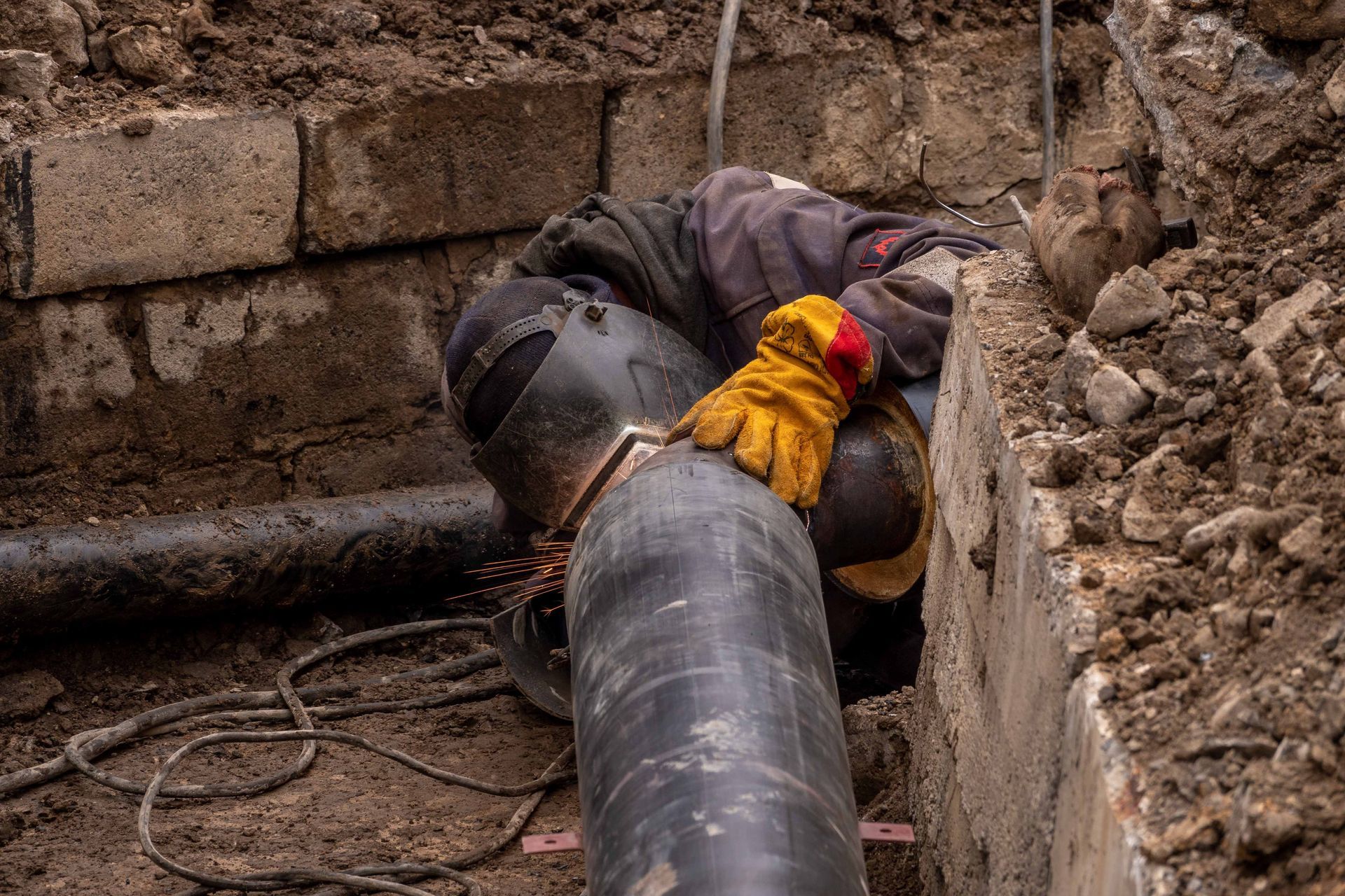 Worker in yellow gloves repairs a large pipe in a muddy trench beside a brick wall.