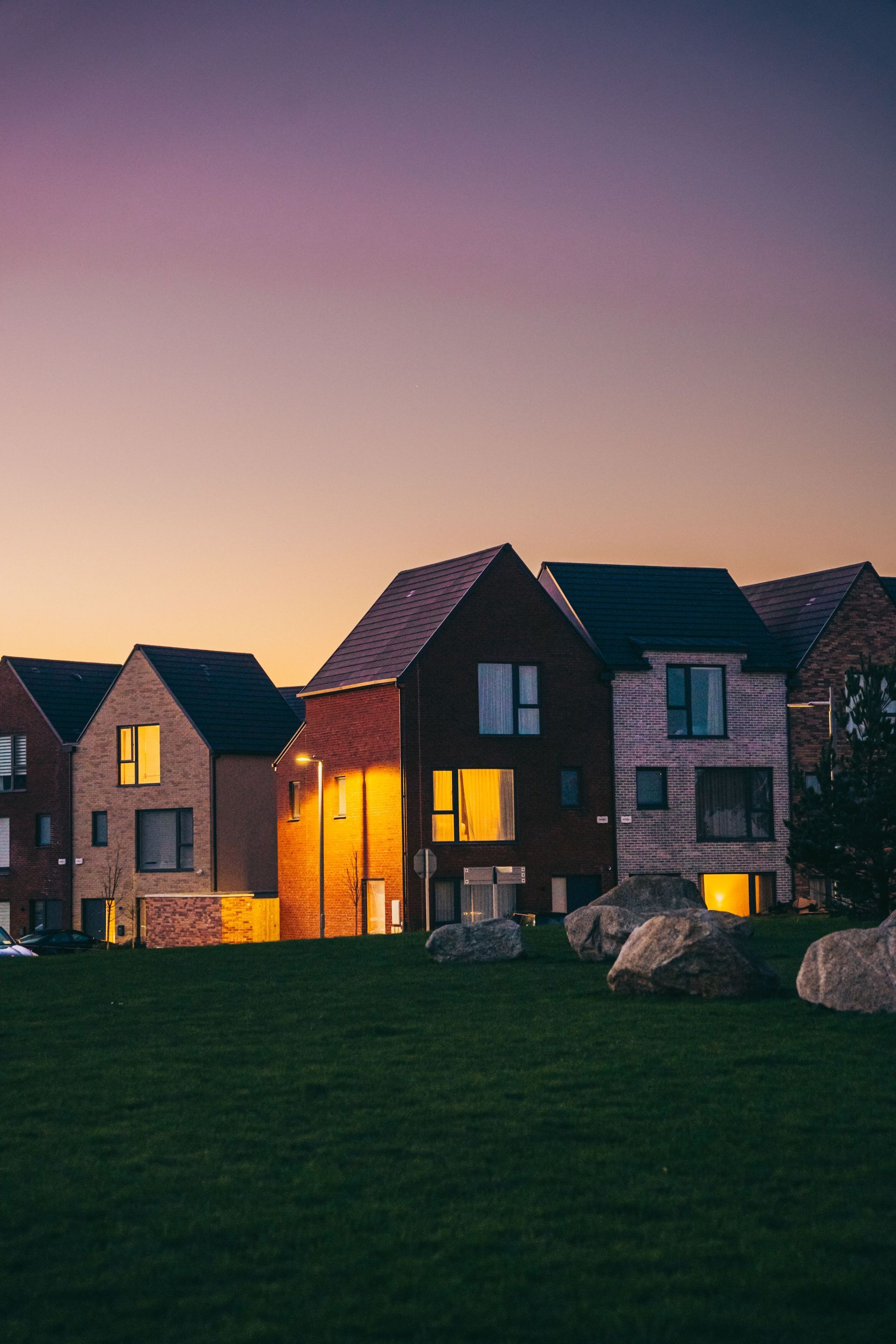 Brick houses at dusk with warm lit windows beside a dark green lawn under a pink-purple sky