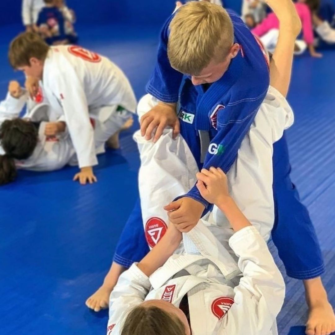 Two people grappling on a mat, one in a black gi, the other in a rash guard, legs entangled.