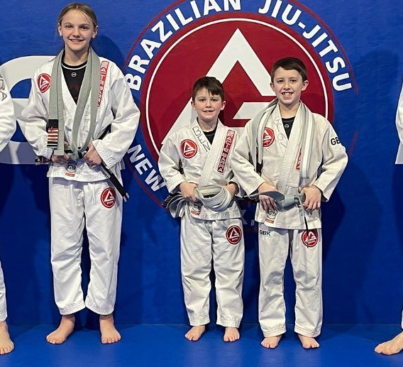 Two young children in blue and black martial arts uniforms grappling on a gym floor, one smiling.