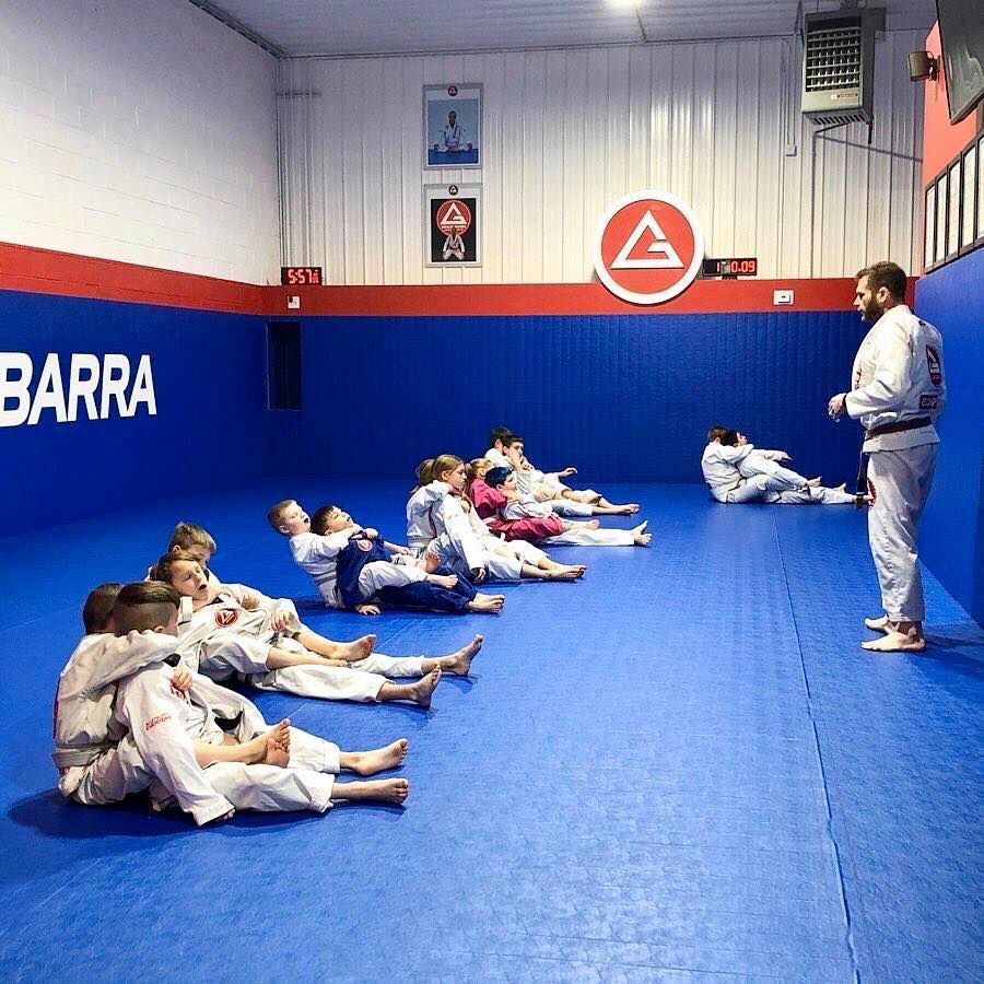 Two children in black and white martial arts uniforms practice a chokehold on a mat. One child smiles.