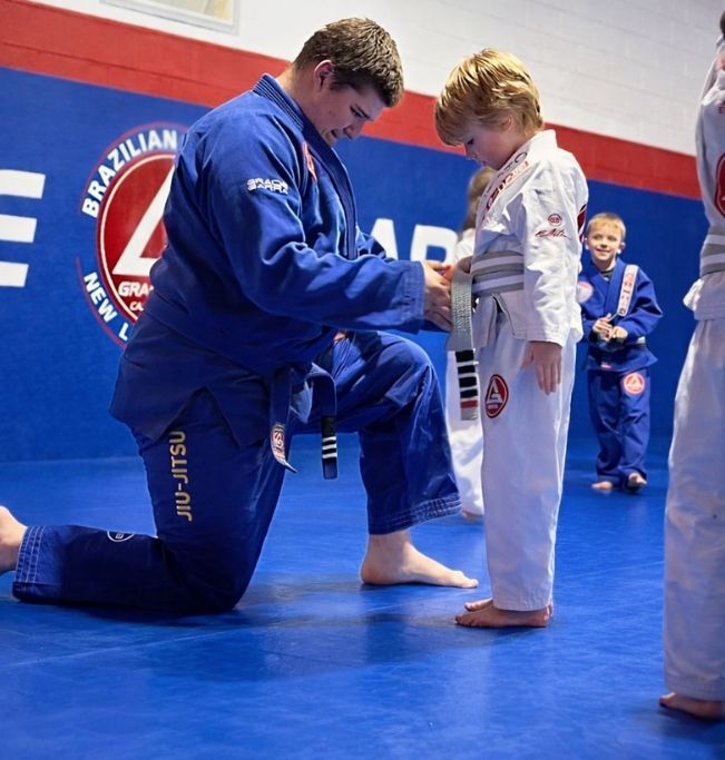 Family in Jiu-Jitsu gis poses. Indoors, blue mats, a smiling woman in the middle, around a seated man with a beard.