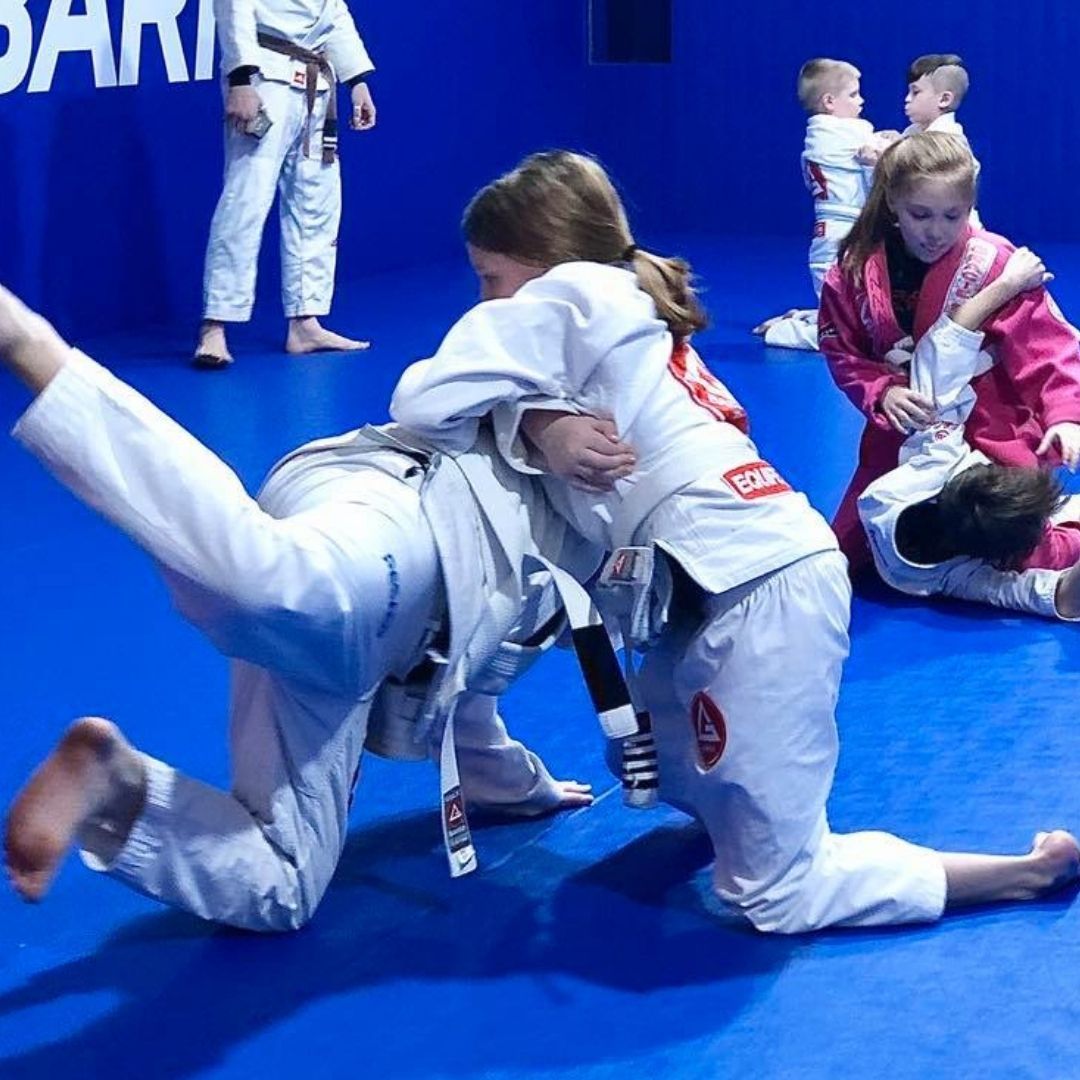 Children in colorful gis play a game on a mat. Two instructors pull a student in blue.
