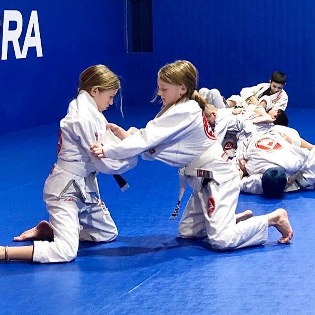 Two children in black and white martial arts uniforms practice a chokehold on a mat. One child smiles.
