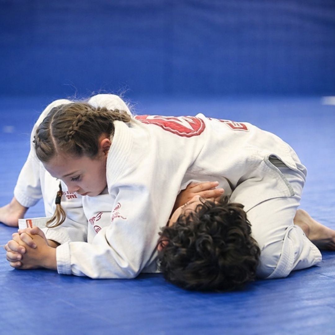 Two children practicing Brazilian Jiu-Jitsu on a mat. One child in white gi has the other in a leg lock. A girl watches.